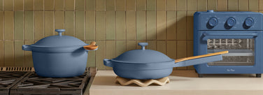 Two blue cookware pieces, a pot with a wooden spoon and a pan with a wooden handle, sit on a kitchen counter next to a blue toaster oven and a gas stove.