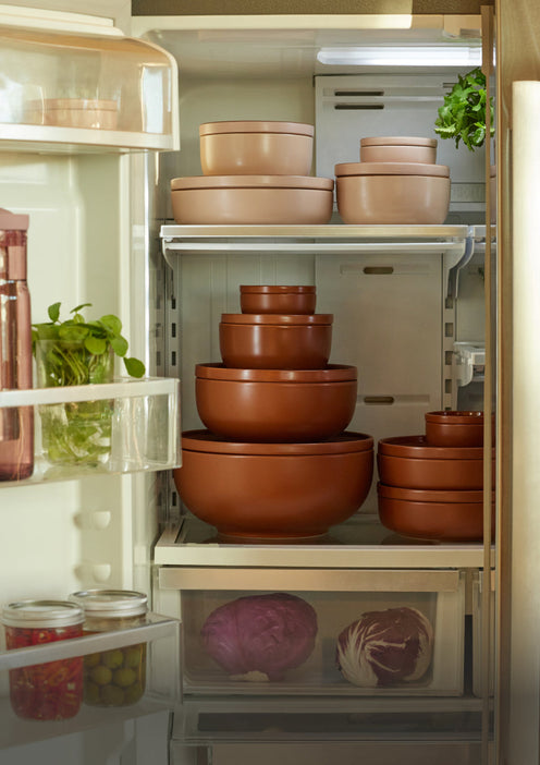 A refrigerator filled with stacked brown and beige food storage containers, jars, leafy greens, and vegetables in the drawers.