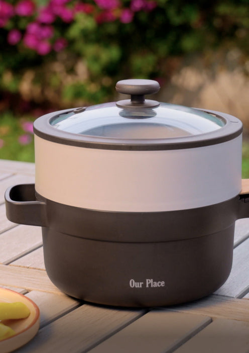 A two-tone cooking pot with a glass lid labeled Our Place sits on a wooden outdoor table, with blurred flowers and greenery in the background.