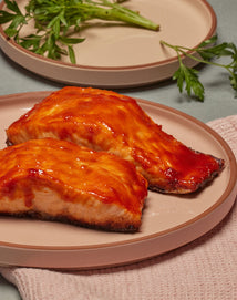 Two pieces of glazed salmon are served on a pink plate, with a beige plate and fresh herbs visible in the background.