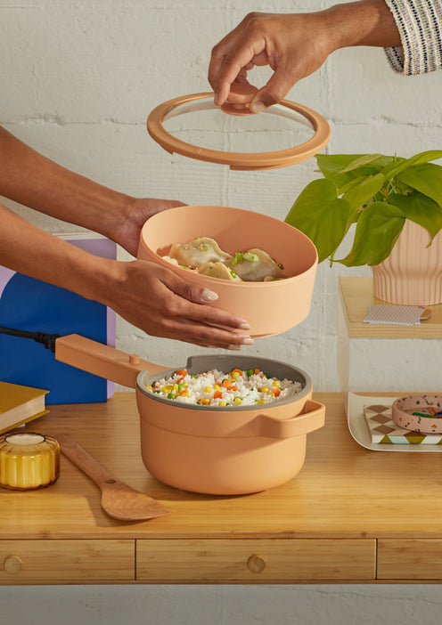 Two hands place a steaming basket with dumplings over a pot of rice and vegetables on a wooden table with books, a plant, and other items.