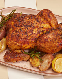 A roasted whole chicken seasoned with spices, garnished with sprigs of rosemary, and served with roasted potatoes on a platter.