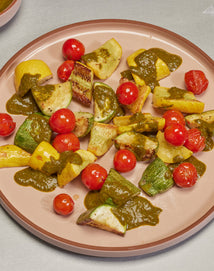 A plate of roasted cherry tomatoes and chopped squash topped with a green sauce, arranged on a pink dish.