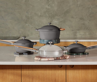 Four modern pots and pans, including gray and stainless steel cookware with wooden handles, are arranged on a kitchen counter with stone trivets.