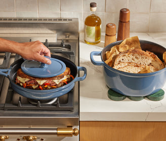 A hand places a lid on a blue Dutch oven containing a sandwich on a stove; next to it is a similar pot filled with sliced bread on a kitchen counter.