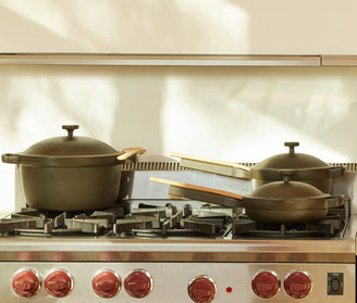 Four green pots and pans with wooden handles sit on a stainless steel gas stove with red knobs in a sunlit kitchen.