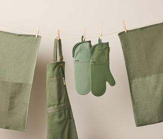A green apron, oven mitts, and two green kitchen towels hang on a clothesline against a neutral background.