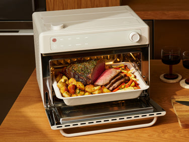 A beige countertop oven with a baking dish containing a roast and vegetables inside.