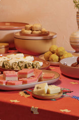 A variety of Indian sweets, including barfi, ladoo, and rolls, are arranged on plates and bowls atop a red festive tablecloth.