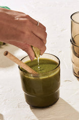 A hand squeezes a lime wedge into a glass of green juice with a wooden spoon inside; two stacked glasses are visible in the background.