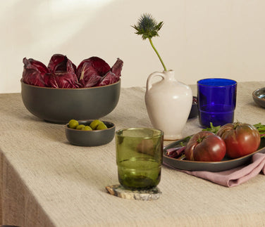 A table set with bowls of vegetables, olives, cheese, crackers, dates, glassware, ceramic vases, and napkins on a neutral tablecloth.