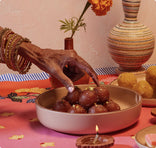 A hand wearing red bangles reaches for a gulab jamun sweet in a bowl, with other sweets, a lit oil lamp, flowers, and pottery on a decorated table.