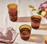 Four amber-colored drinking glasses, two stacked, on a light tablecloth with a pink textured cloth, citrus fruits, and fruit peels nearby.