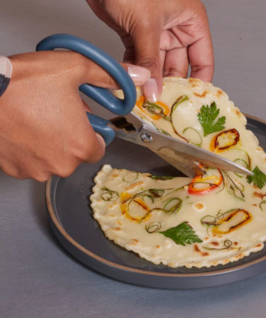 A hand uses scissors to cut a flatbread with herbs and vegetables on a round gray plate.
