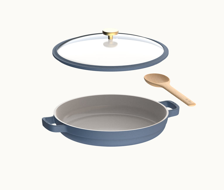 A blue ceramic baking dish with handles, a transparent glass lid with a gold knob, and a wooden spoon, all shown on a plain background.
