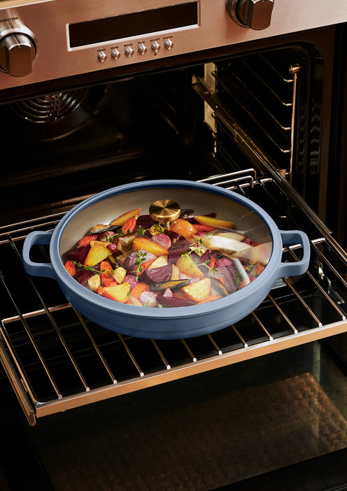A blue covered roasting pan with vegetables and herbs sits on an oven rack inside a stainless steel oven.