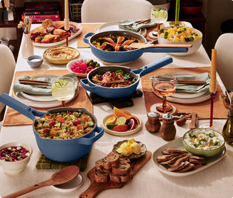 A dining table set for a meal features various dishes in blue cookware, salads, bread, drinks, and tableware arranged neatly on placemats with candles and condiments.