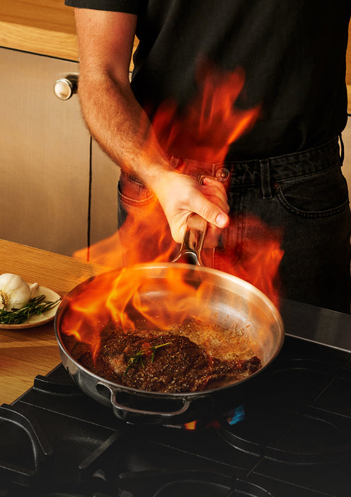 A person searing a steak in a flaming pan on a stovetop.