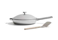 A white lidded frying pan with a long handle sits next to a metal spatula with a beige handle, both on a white background.
