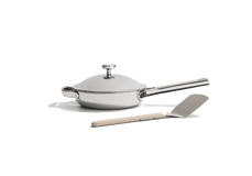 A stainless steel frying pan with a lid is placed next to a spatula with a beige handle on a white background.