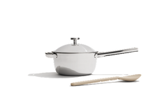 A stainless steel saucepan with a lid and a long handle is placed beside a beige cooking spoon on a white background.