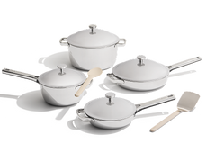 A set of four stainless steel cookware pieces with lids, a wooden spoon, and a spatula arranged on a white surface.