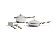 Two stainless steel pans with lids, a metal spatula, and a wooden spoon are arranged on a white background.