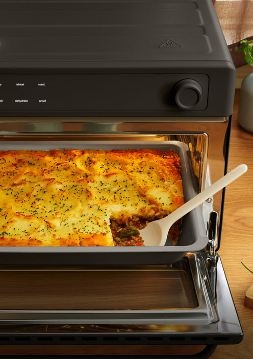 Shepherds pie in a baking tray inside an open countertop oven, with a serving spoon scooping out a portion.