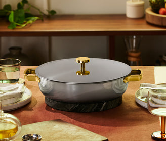 A gray cooking pot with gold handles and lid sits on a marble trivet on a set dining table with plates, glasses, and napkins.