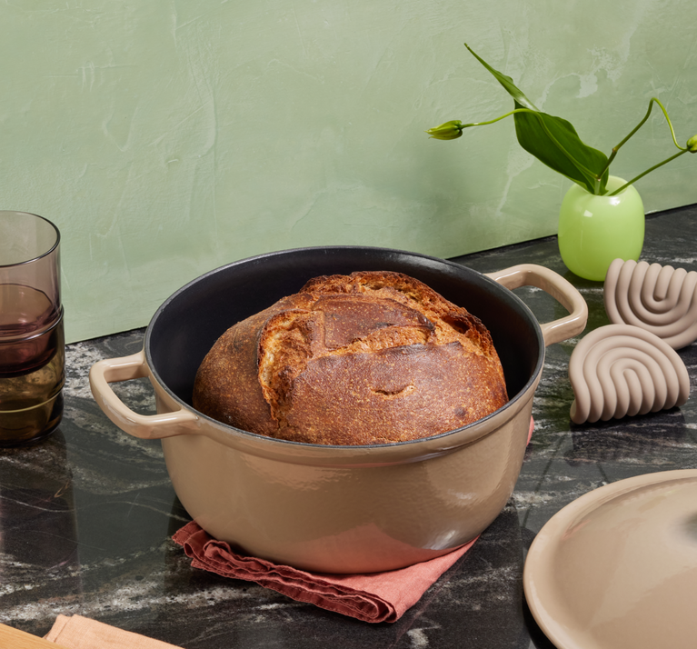 A round loaf of bread sits in a beige Dutch oven on a dark marble countertop, with a green vase, two glasses, pot holders, and the pot lid nearby.