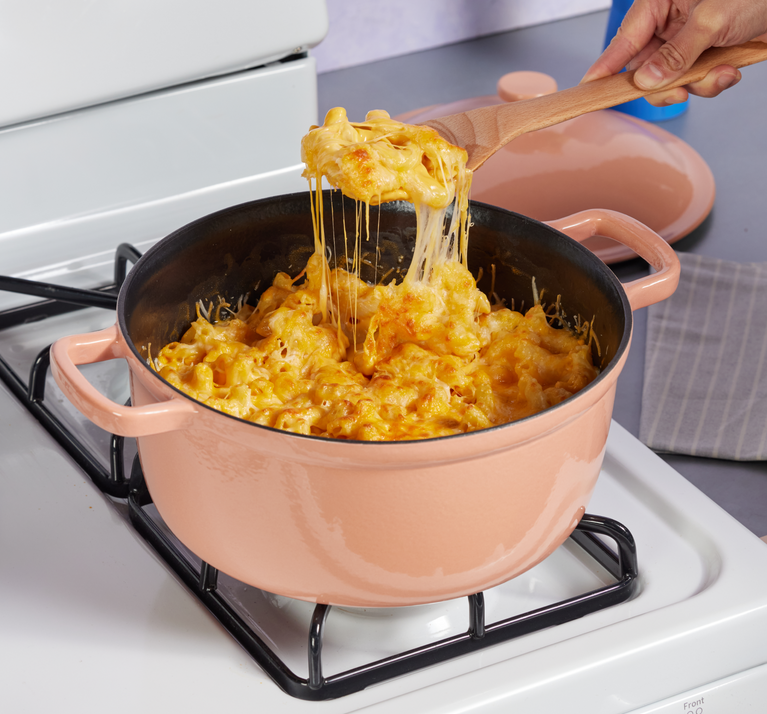 A person lifts a wooden spoonful of cheesy macaroni from a pink pot on a stovetop.