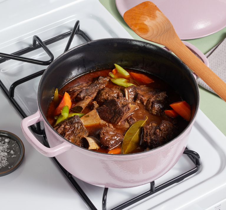 A pink Dutch oven on a stove contains beef stew with carrots, celery, potatoes, and bay leaves. A wooden spoon and a lid rest nearby.