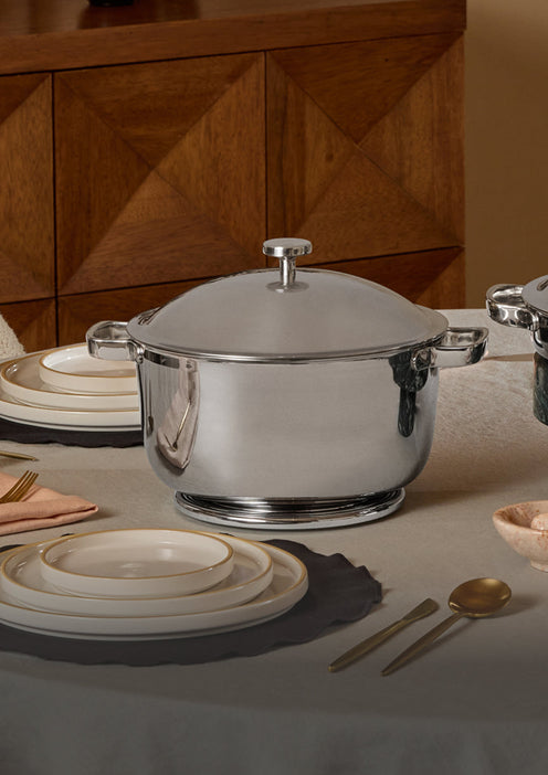 A shiny stainless steel pot with a lid is placed on a round, covered dining table set with white plates, gold utensils, and a napkin. In the background is a wooden cabinet with geometric patterns.