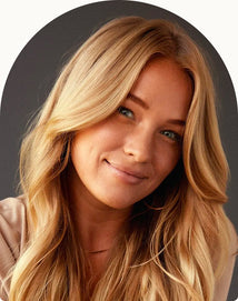 A woman with long, wavy blonde hair and light skin is smiling softly at the camera against a dark background.