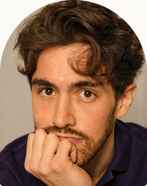 A man with wavy brown hair and a trimmed beard rests his chin on his hand, looking directly at the camera with a thoughtful expression.