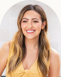 A woman with long, wavy, light brown hair wearing a yellow sleeveless top smiles at the camera against a light background.