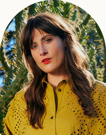 A woman with long brown hair wearing a yellow eyelet shirt stands outdoors in front of a cactus, looking at the camera.