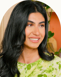 A woman with long black hair wearing a green floral top smiles and looks to the side. There are green plants in the background.