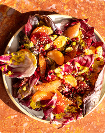 A plate of radicchio salad topped with orange segments, pomegranate seeds, pistachios, and a yellow dressing on a marbled orange surface.