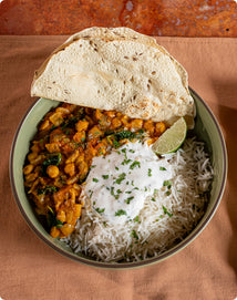 A bowl of basmati rice topped with vegetable curry, yogurt, chopped herbs, a lime wedge, and a piece of papadum on the side.