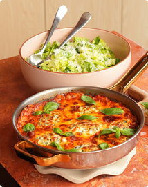 A baked dish topped with melted cheese and basil sits in a pan next to a bowl of green salad with shredded cheese and two serving utensils.
