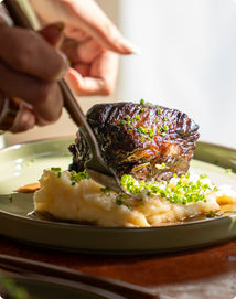 A hand cutting into a piece of braised meat served on mashed potatoes and garnished with chopped chives on a green plate.