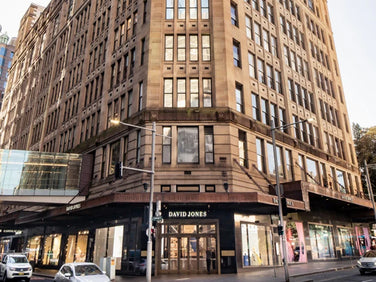 A large multi-story sandstone building with a David Jones store on the ground floor, located on a busy city street corner with cars and pedestrians.