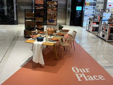 Display of Our Place cookware and tableware on a wooden dining table with chairs, set up in a store with various home goods on shelves in the background.