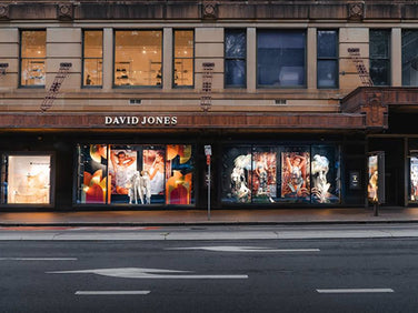 Street view of a David Jones store with large, illuminated window displays featuring decorative installations and mannequins; the storefront appears quiet with no visible pedestrians.
