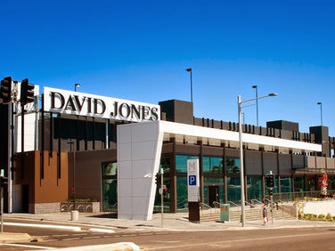 Modern David Jones department store exterior with large signage, glass entrance, and adjacent street with parking signs under a clear blue sky.
