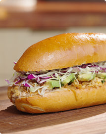 A sandwich on a wooden surface, filled with shredded chicken, sliced avocado, and shredded cabbage inside a shiny toasted roll.
