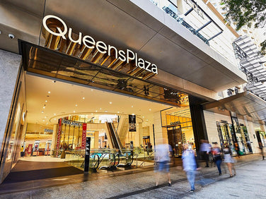 Entrance to QueensPlaza shopping mall with glass doors, visible escalators, and several blurred people walking in and out.