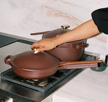A person lifts the lid of a brown skillet on a gas stove, with a matching pot and white marble backsplash in the background.