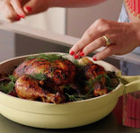 Person seasoning roasted chicken in a pan with herbs